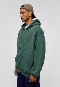 Groene rits hoodie met een voorzak, trekkoordkap en geribbelde manchetten, gecombineerd met een tan kleurige baseballcap. Gemaakt van zacht stof, met een ontspannen pasvorm.