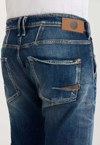 Jeans de denim en azul oscuro con ligero desgaste, que presentan múltiples bolsillos y una etiqueta de cuero marrón en la cinturilla.