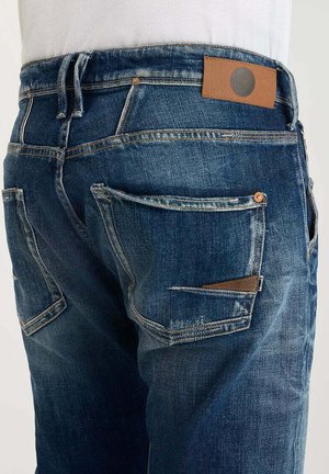 Donkerblauwe denim jeans met lichte slijtage, voorzien van meerdere zakken en een bruine leren patch op de tailleband.