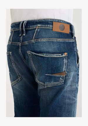 Donkerblauwe denim jeans met lichte slijtage, voorzien van meerdere zakken en een bruine leren patch op de tailleband.