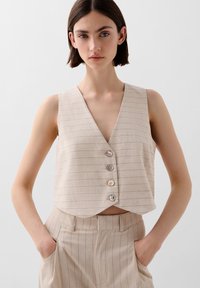 Beige cropped vest met een diepe V-hals, voorzien van horizontale strepen en vijf iriserende knopen. Gepaard met bijpassende gestreepte broek.
