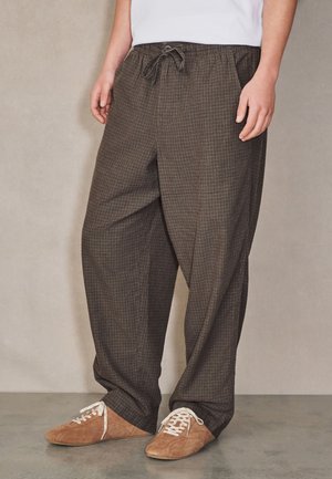 Person trägt lockere braune kariert Hose mit Kordelzug in der Taille und hellbraune Wildleder-Schnürschuhe, steht vor neutralem Hintergrund.