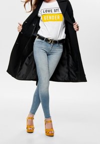Abrigo largo negro sobre una camiseta gráfica blanca con texto amarillo y negro. Jeans ajustados de color azul claro y tacones de plataforma amarillos completan el conjunto.