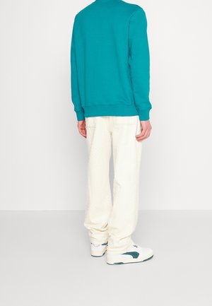 Un sweat-shirt teal avec des poignets côtelés, associé à un pantalon blanc texturé. Les chaussures présentent une base blanche avec des accents verts, vues de l'arrière.