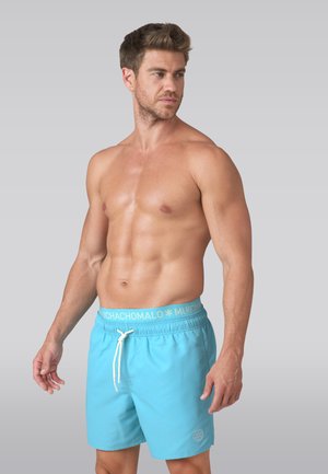 Muţ svalnatý muž bez trička, oblečený v svetlomodrých plavkových šortkách s bielym šnúrkou, pozerajúci sa doľava na sivom pozadí.