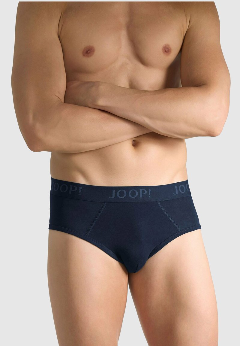 Mężczyzna o umięśnionej sylwetce z założonymi skrzyżowanymi ramionami, ubrany w ciemnoniebieskie majtki JOOP! na jednolitym jasnoszarym tle.