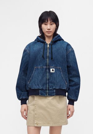 Giovane che indossa una felpa con cappuccio in denim blu con zip e tasca frontale e una gonna beige al ginocchio su uno sfondo bianco semplice.