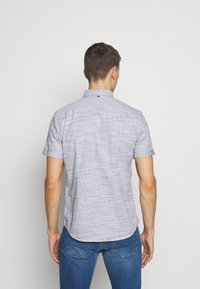 Camisa de cuadros azul claro de manga corta con cuello abotonado y una textura sutil. Presenta un pequeño logo en la parte trasera del cuello.