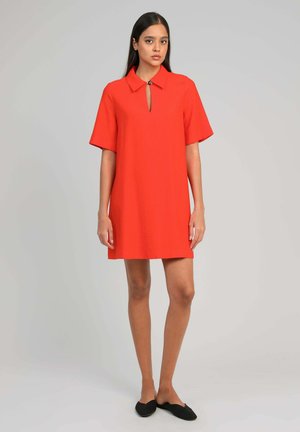 Femme debout portant une robe rouge à manches courtes, longueur genou, avec un col et un détail de bouton en forme de trou de serrure, associée à des chaussures noires à enfiler.