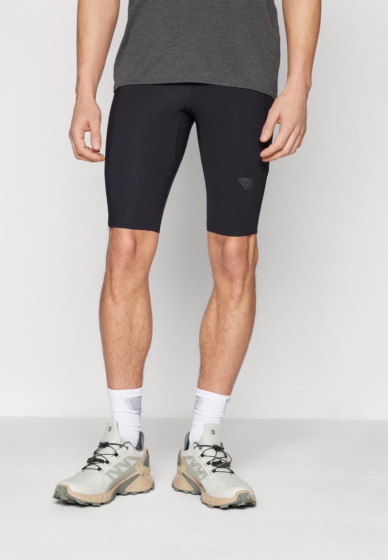 Black cycling shorts in stretchy material, featuring a fitted design and a small logo on the side. Paired with grey and beige athletic shoes.
