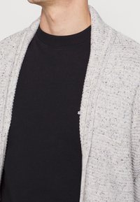 Světle šedý pletený cardigan s texturovaným vzorem, otevřeným předním dílem a šálovým límcem, nošený přes černé tričko s kulatým výstřihem.