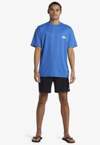 Modrá tričko s krátkým rukávem a kulatým výstřihem, černé kraťasy. Výrazné logo Quiksilver na levé části hrudi. Nosené s žabkami.