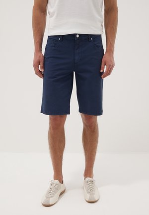Mann, der marineblaue knielange Shorts, ein weißes T-Shirt und weiße Turnschuhe trägt, steht vor einem einfarbigen hellen Hintergrund.