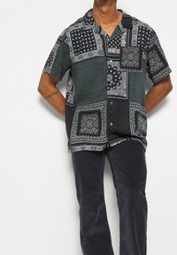 Camisetta nera a maniche corte con dettagli paisley e decorativi, dotata di taschino sul petto e chiusura con bottoni. Indossata con pantaloni scuri.