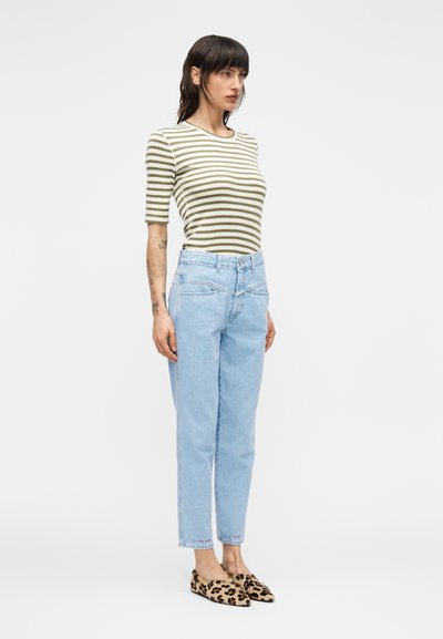 Γυναίκα με σκουρόχρωμα μαλλιά, που φοράει ριγέ πουκάμισο σε ελιά και λευκό, ανοιχτό μπλε τζιν ψηλόμεσο και παπούτσια τύπου slip-on με λεοπάρ σχέδιο, στέκεται μπροστά σε λευκό φόντο.