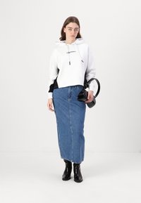 Un hanorac scurt alb și negru cu logo, asortat cu o fustă lungă din denim albastru și cizme negre până la gleznă. Ținând o geantă mică neagră.