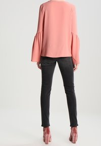 Femme vue de dos portant un chemisier rose ample à manches évasées, un jean skinny noir et des chaussures à talons carrés en velours rose.