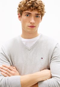 Junger Mann mit lockigem blondem Haar und blauen Augen, der einen hellgrauen V-Ausschnitt-Pullover über einem weißen Hemd trägt und mit verschränkten Armen steht.