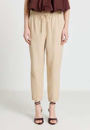 Femme portant un pantalon beige à cordon avec poches avant et poignets élastiques, associé à des sandales à talons à brides marron foncé.