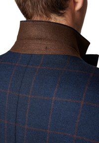 Kołnierz wełnianej marynarki z brązowym filcowym spodem, zdobiony niebieską kratą z subtelnymi brązowymi akcentami. Strukturalny fason i teksturowany materiał.