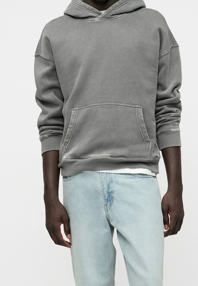 Homme portant un sweat à capuche gris délavé avec poche avant et un jean en denim bleu clair, debout devant un fond uni.