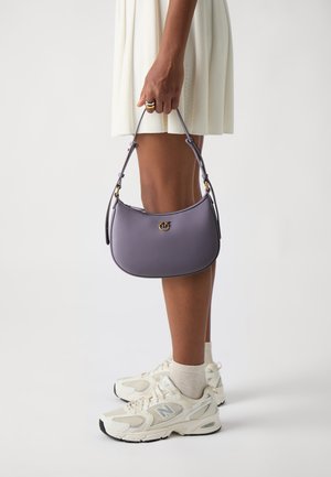 Bolso de piel púrpura con forma curvada, detalles en dorado y una correa delgada ajustable. Exhibido junto a unas zapatillas blancas y una falda clara.