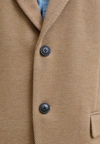 Brauner strukturierten Blazer mit Reverskragen, zwei schwarzen Knöpfen und einer Brusttasche. Der Stoff wirkt gewebt und strapazierfähig.