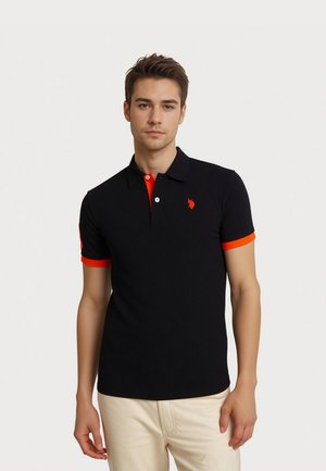 Jeune homme portant un polo noir avec des bordures rouges et un pantalon beige, debout devant un fond neutre.