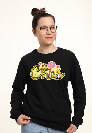 Kvinde med briller og håret i en knold iført en sort sweatshirt med gulternet "Cher" tekst og pink hjerte og pompom grafik.
