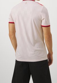 Mies, który ma na sobie jasnoróżową koszulkę polo z krótkim rękawem z czerwonymi wykończeniami na rękawach i kołnierzyku, w zestawieniu z czarnymi spodniami, pokazany od tyłu.