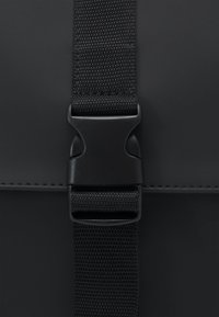 Black bag with a textured, matte finish. Features a black plastic buckle and a woven strap, providing a sleek, minimal design.