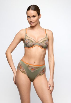 Frau trägt ein olivgrünes Dessous-Set mit orangefarbener Einfassung und floralen Spitzen-Details, posiert vor weißem Hintergrund.
