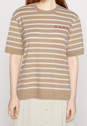 Beige og hvidt kortærmet skjorte med vandrette striber og rødt "GANT"-logo på brystet båret af en person med lys hud og langt hår.