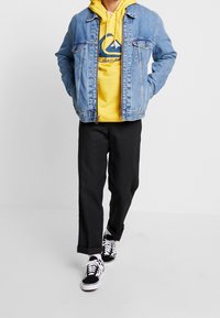 Jeansjacka med ljus tvätt, gul huvtröja med logotyp, svarta raka byxor och svarta med vita mönstrade sneakers.