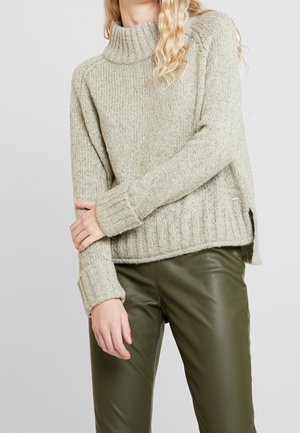 Persoon draagt een lichtgrijze gebreide coltrui en olijfgroene leren broek, met de armen over de borst gekruist.