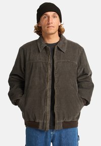 Giacca in corduroy di colore grigio scuro, con zip frontale, polsini e colletto a coste, tasche laterali e un sottile logo del marchio sul petto.