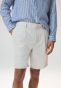 Shorts en lin gris clair avec plis avant, poches latérales et texture lisse, associée à une chemise à manches longues rayée bleue.