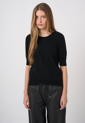 Young woman with long blonde hair wearing a black short-sleeve sweater and black leather pants standing against a plain background.