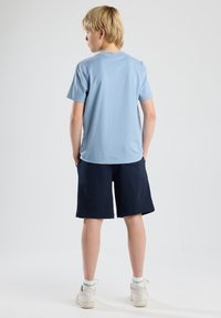 Vaaleansininen puuvillapaita ja tummansiniset shortsit, molemmat rentoja tyylejä. Paidassa on lyhyet hihat, kun taas shortseissa on sileä rakenne. Valkoiset lenkkarit.