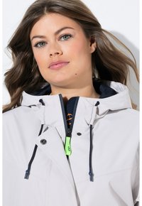 Veste gris clair avec capuche, doublure bleu marine, fermeture éclair à l'avant et cordons de serrage. Présente un tire-zipp vert et des boutons noirs pour la fermeture.
