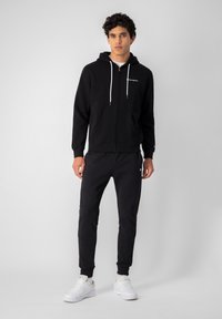 Czarna bluza z kapturem na zamek z białymi sznurkami, w połączeniu z czarnymi spodniami dresowymi z białymi akcentami z logo. Model nosi białe sneakersy.
