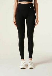 Černé žebrované legíny s vysokým pasem a logem, které mají hladkou texturu a obepínající siluetu. Perfektně se hodí k sportovní obuvi.