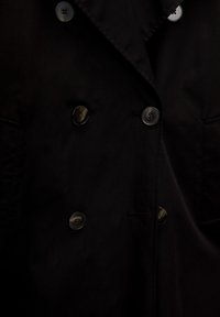 Manteau noir croisé avec de gros boutons, deux poches avant, et une texture de tissu lisse et mate. Doté d'un col à revers.