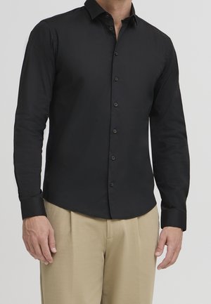 Schwarzes Langarmhemd aus glattem Stoff, mit schmaler Passform, Button-Down-Kragen und schwarzen Knöpfen. Getragen mit beigen Hosen.