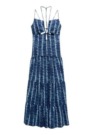 Frau steht in einem ärmellosen Maxi-Kleid mit blauer Batikmusterung, mehrlagigem Rockteil, und trägt schwarze flache Sandalen mit Knöchelriemen.