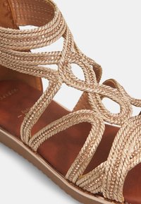 Kultainen letitetty sandaali kimaltelevilla aksenteilla, jossa on pehmeä ruskea pohja ja monimutkainen ristikkäinen muotoilu. Tasapohja mukavuutta varten.