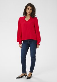 Blouse rouge à col en V avec manches longues bouffantes, associée à un jean skinny bleu foncé et des escarpins noirs à bride arrière. Tissu lisse, coupe décontractée.