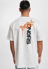 Balta medvilnė t-shirt su per dideliu pjūviu. Ant nugaros turi drąsų oranžinės ir juodos spalvos grafikos tekstą. Trumpomis rankovėmis, apvalia iškirpte.