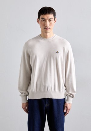 Hombre vistiendo un suéter de manga larga beige claro con un pequeño logotipo colorido en el pecho y pantalones azul oscuro, de pie contra un fondo liso.