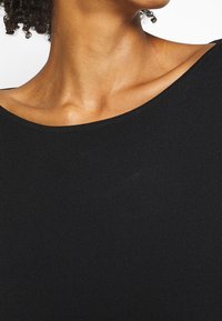 Débardeur noir à côtes avec un col large et une texture lisse. Une vue en gros plan met en évidence l'élasticité et la douceur du tissu.